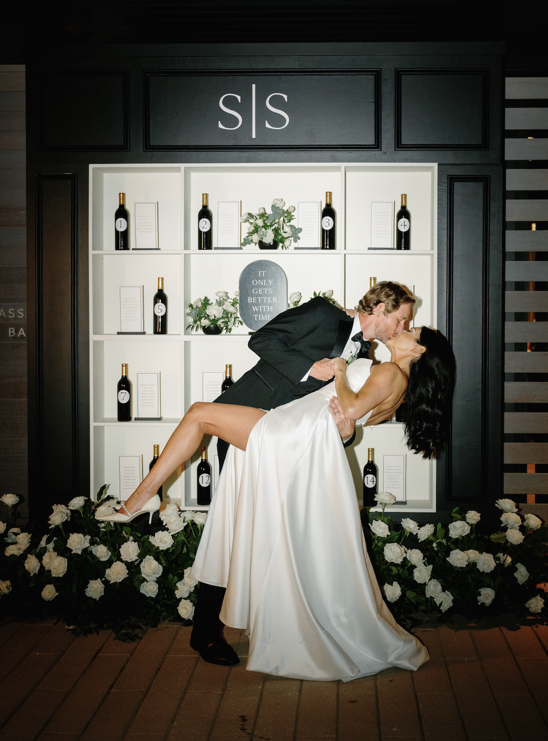 A couple kisses in front of a display shelf with wine bottles and flowers, underneath a monogram "S|S." The woman is wearing a white gown, and the setting suggests an elegant event.